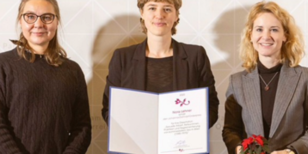 Drei Frauen stehend und in die Kamera blickend. Nora Lehner in der Mitte, hält Verleihungsurkunde in die Kamera