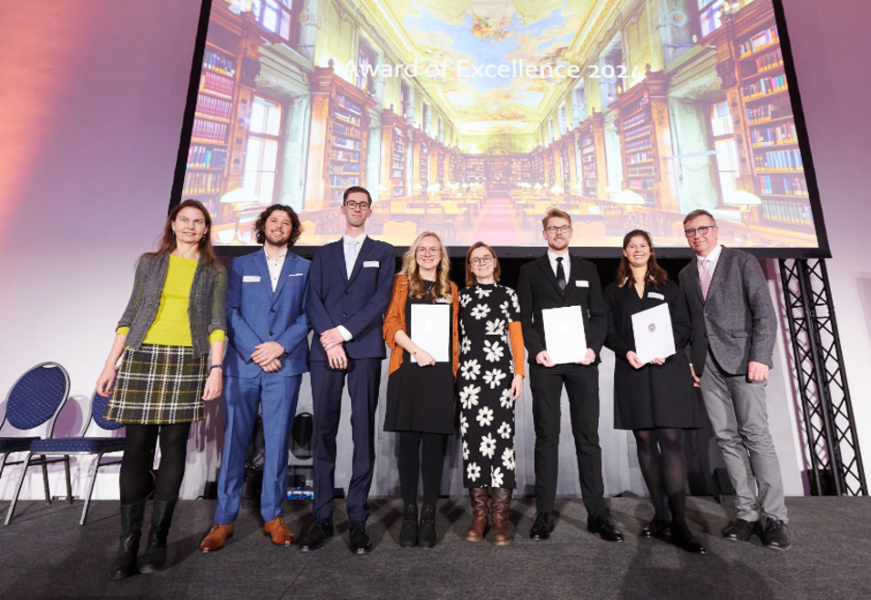 Preisträger:innen der Universität Wien des Award of Excellence 2024 mit Urkunden vor einem Screen stehen